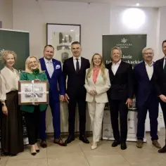 Grupa osób w eleganckich strojach, wśród nich kurator, pozuje do zdjęcia. Jedna z pań trzyma w rękach oprawiony obraz.