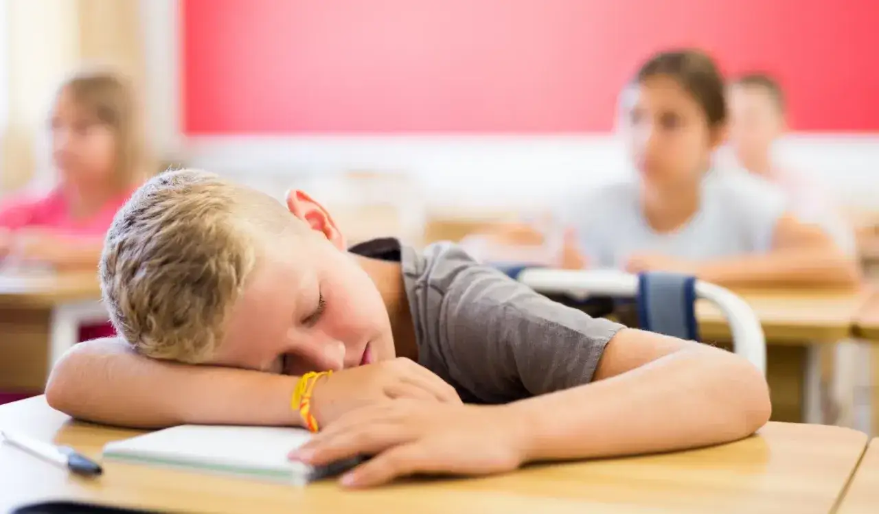 Chłopiec śpi na lekcji, jego głowa oparta na rękach. W tle widać innych uczniów, tworząc dysonans między aktywnością a odpoczynkiem.