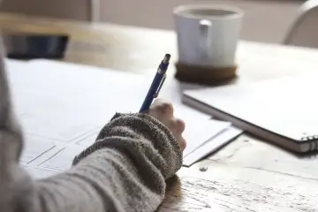 Ręka pisze długopisem na papierze, tworząc szkic. Obok leży notatnik i kubek kawy. Tak wygląda referat w trakcie tworzenia.