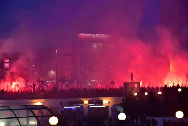 Tłum z polskimi flagami i racami na tle nocnego miasta. Widać billboard Santander. Manifestacja, która może być wyrazem etnocentryzmu.